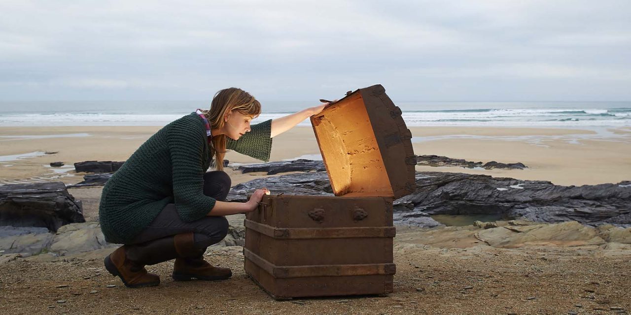 A woman looks inside a treasure chest with gold on a deserted beach.